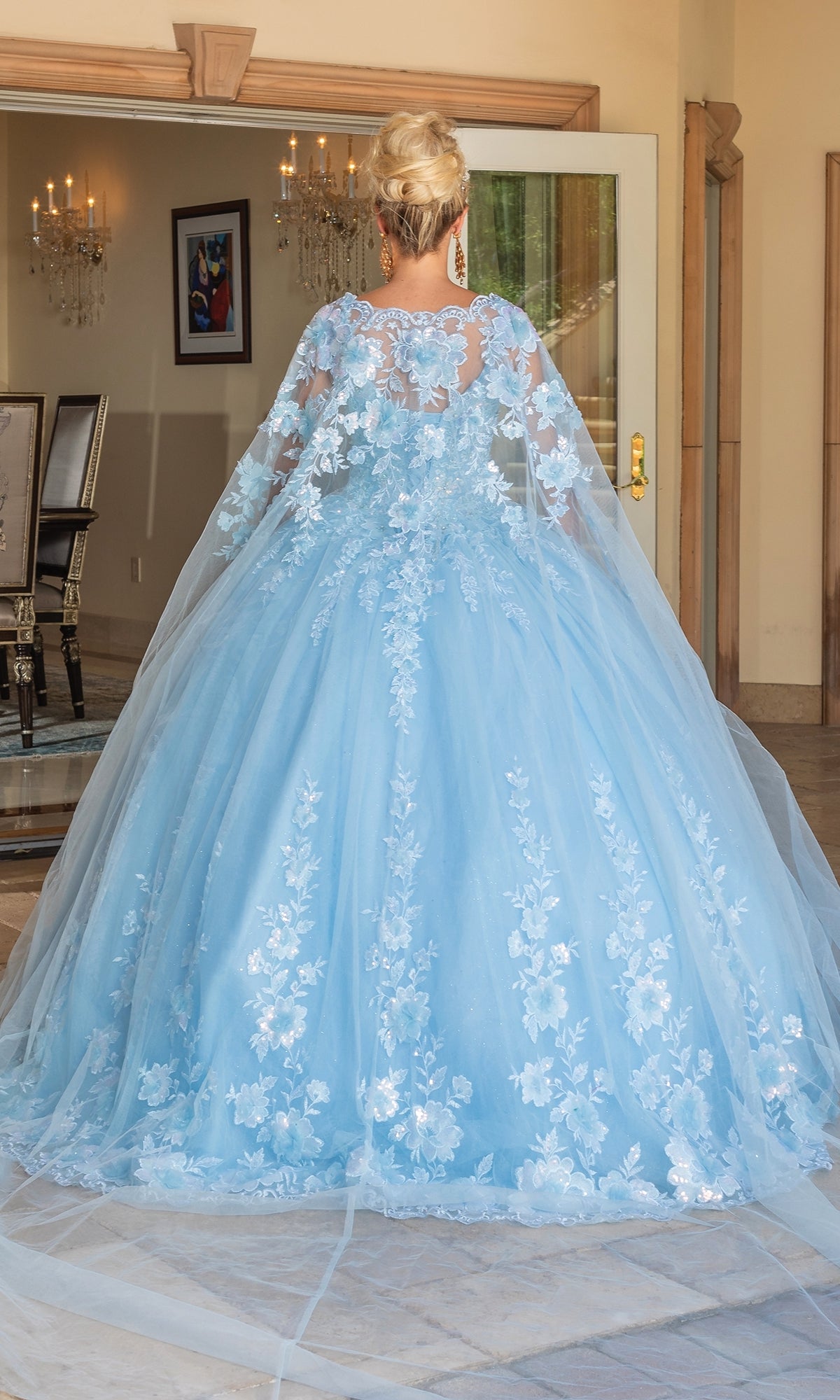 A person with blonde hair in an updo, seen from behind indoors, is wearing the Quinceanera Dress 1778 By Dancing Queen—a light blue ball gown featuring floral lace details and a lace-embellished cape.